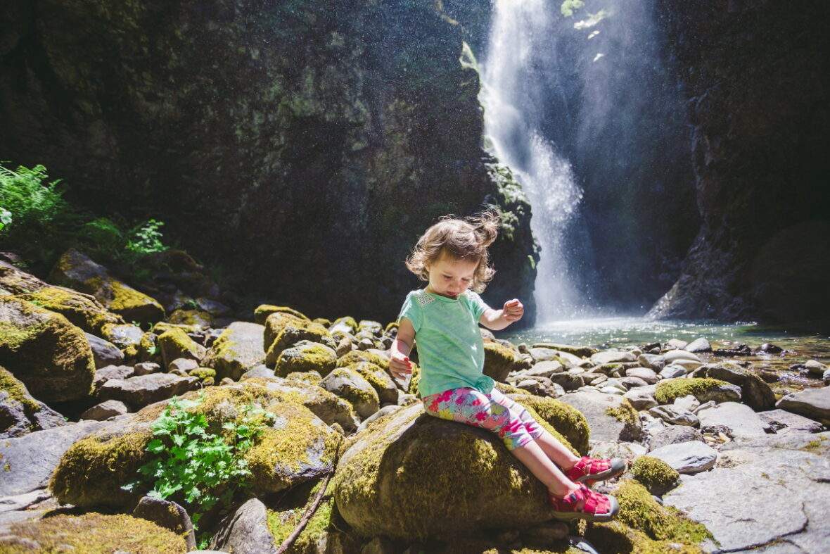 Little girl sitting on mossy rock in front of waterfall in Oregon